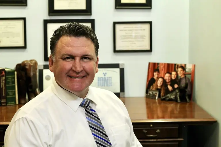 Dr. Raymond Espinosa smiling at his desk in a white shirt and striped tie, with framed diplomas and a family photo behind him